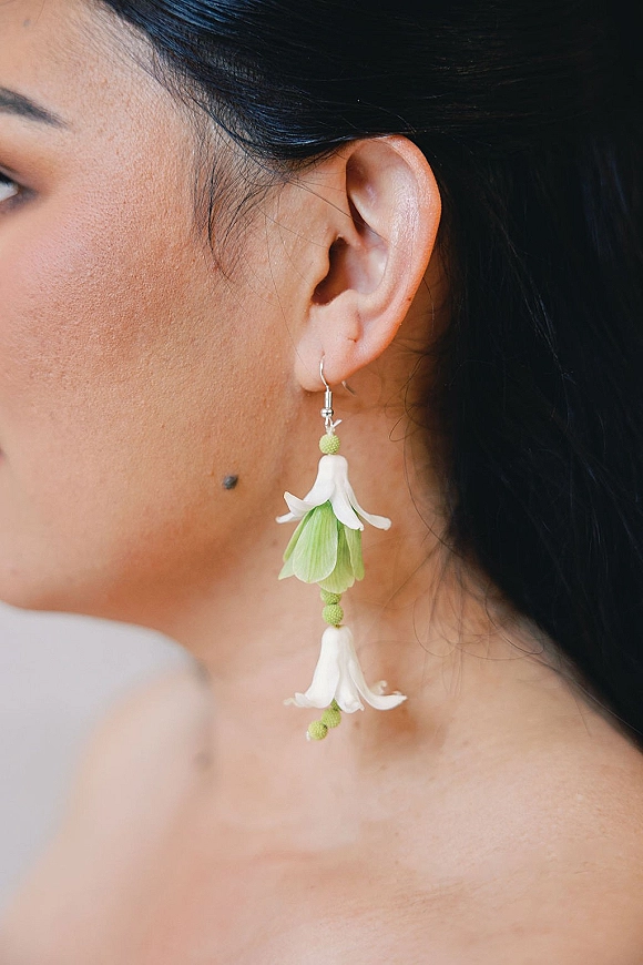 Bridal earrings with white flower petals and green floral beads dangling from an ear hook on a bride’s side profile against dark hair