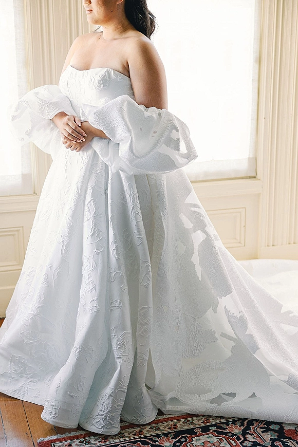 Wedding dress with a strapless wedding dress bodice and off-the-shoulder sleeves, textured ball gown skirt and train by a sunlit window