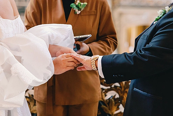 Wedding ring exchange close up as groom slides a gold band onto the bride’s finger, suit cuff and wristwatch beside officiant in indoor ceremony