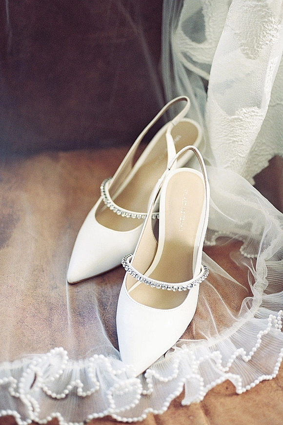 Bridal shoes, white wedding heels with rhinestone ankle straps beside a tulle veil with lace trim and pearl edging on a wood floor by a chair