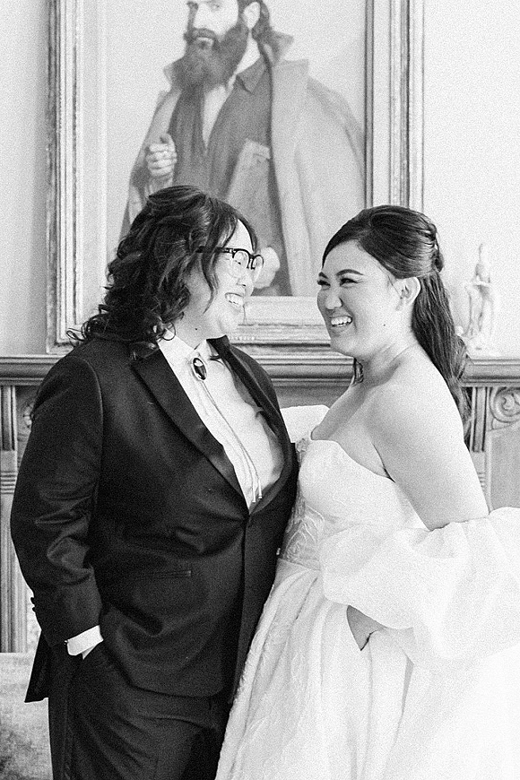 Couple portrait of a lesbian wedding couple laughing and looking at each other, one in strapless dress, indoors by ornate molding and framed painting
