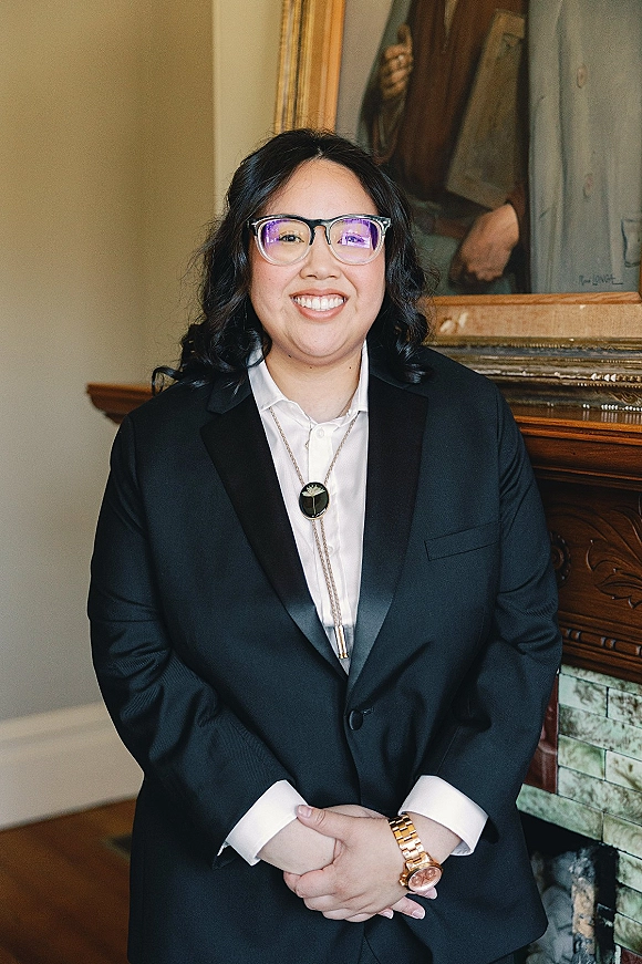 Groom portrait in a black tuxedo with a bolo tie and glasses, standing by a tiled fireplace mantel with framed painting behind