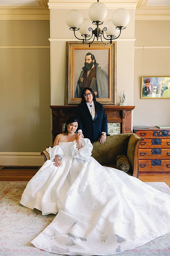 Couple portrait of bride seated on a vintage sofa and groom standing behind her in a living room with fireplace mantel and chandelier