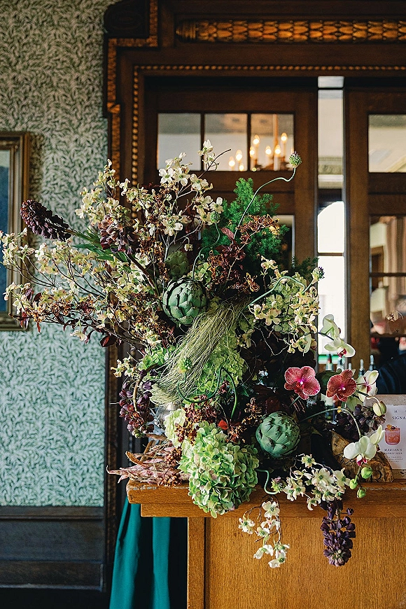 Wedding floral arrangement with orchids and hydrangea cascading from a wooden stand beside a bar menu sign under chandelier lights indoors
