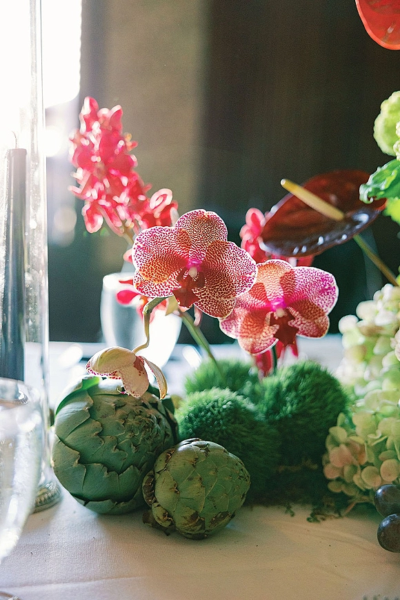 Wedding centerpiece with orchids and hydrangeas, artichokes and greenery beside a glass cylinder candle on a sunlit tabletop against dark backdrop