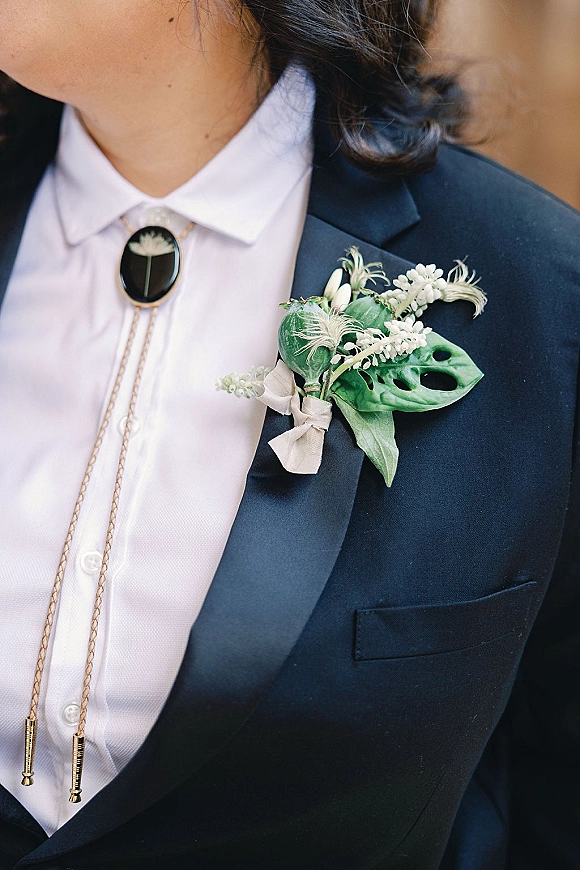 Groom boutonniere with monstera leaf and lily of the valley pinned to a navy suit lapel, paired with a black bolo tie in soft outdoor blur