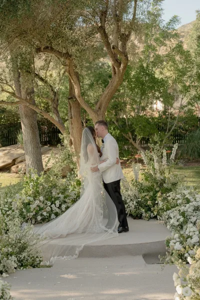 Ceremony kiss moment as the bride and groom embrace, her long train sweeping a tree-lined garden lawn.