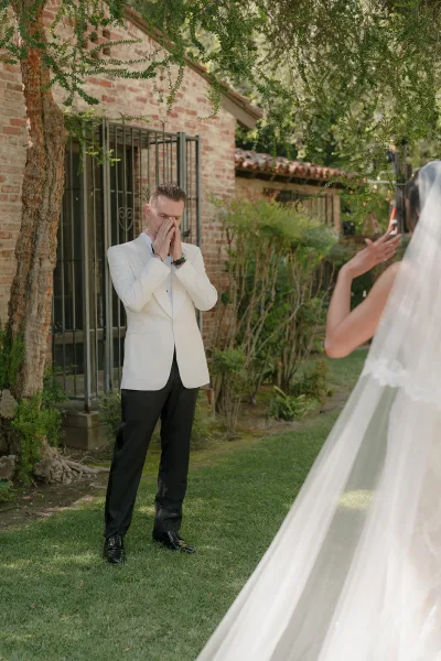 First look moment with tender groom reaction as the bride's long veil frames them by a brick wall and iron gate in a leafy garden.