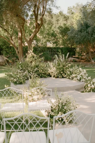 Ceremony setup featuring refined outdoor ceremony decor, with white florals bordering the aisle amid garden trees, grass lawn, and boulders.
