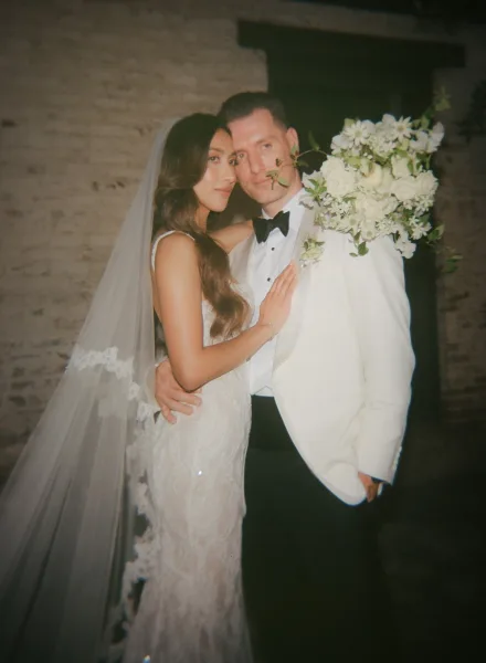 Bride and groom portrait in a tender embrace, she holds a white and green bridal bouquet against a brick wall beside a dark doorway.
