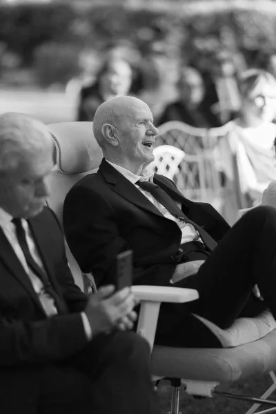 Ceremony moment as a candid wedding guest smiles, holding a smartphone amid seated guests and chairs against lush outdoor greenery.