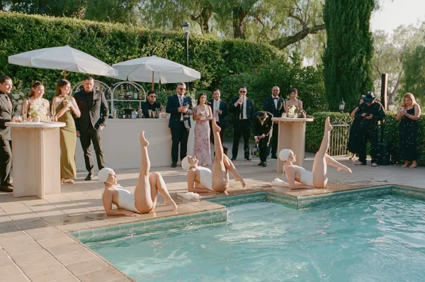 Cocktail hour entertainment centers on a synchronized swimmers wedding show in the pool, performers in shimmering swim caps framed by garden hedges.