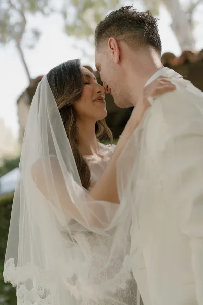 Couple portrait under a lace-trimmed veil, faces nearly touching in a sunlit garden courtyard by a stone wall and clay tile roof.