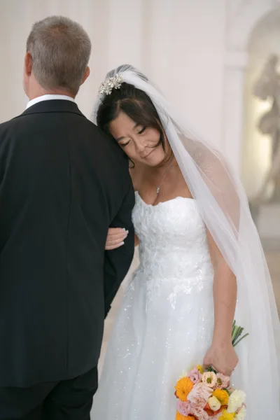 Bride and father walk down the aisle as he gives her away, she holds a pink and orange bouquet with a sparkly veil by a white wall indoors