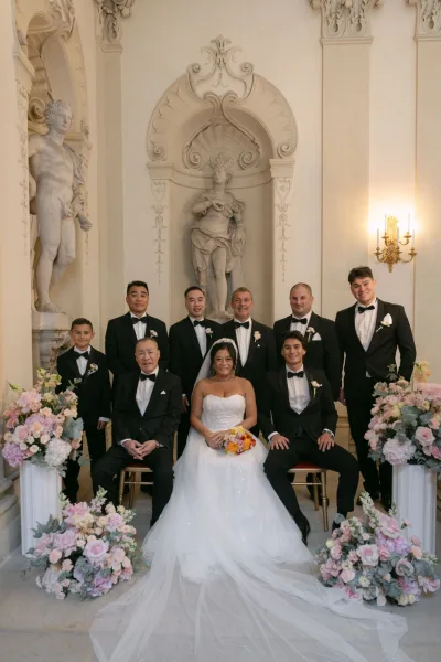 Wedding party portrait with bride and groom with groomsmen, bride in strapless gown holding bouquet in an ornate hall with statues