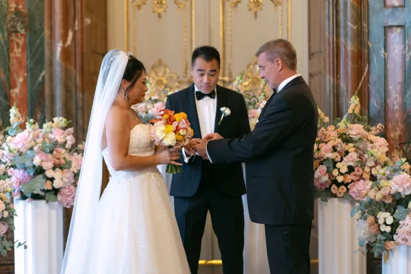 Wedding vows during a ring exchange ceremony as bride and groom hold hands, her veil and bouquet framed by gilded walls and marble columns