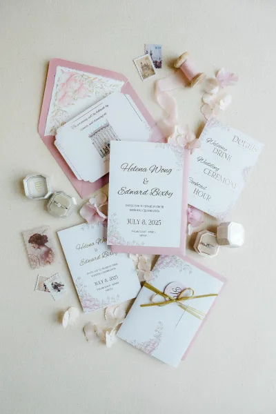 Wedding invitation suite with blush pink wedding invitations, pink envelopes, wax seal, ribbon spool, and rings arranged on a linen backdrop