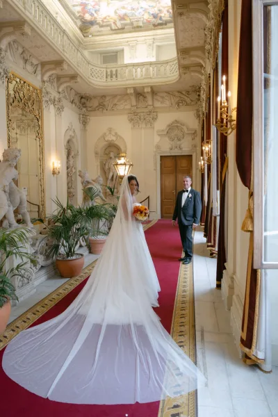 Bride portrait in a cathedral veil, holding an orange bouquet, walking down a red-carpeted ornate ballroom hallway with statues and fresco ceiling