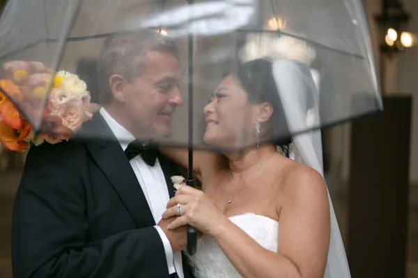 Couple portrait of bride and groom under umbrella, embracing with bouquet and veil at a venue entrance doorway with wall sconces