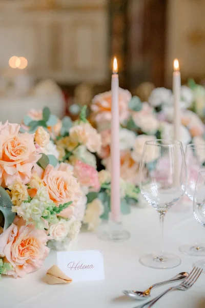 Reception tablescape with a wedding table centerpiece of peach roses and eucalyptus, taper candles, place card, fortune cookie, and wine glasses amid bokeh lights