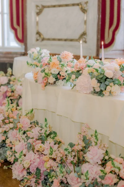Wedding sweetheart table with sweetheart table flowers in blush and peach, hydrangeas and roses, white taper candles, and cascading floor florals against ornate paneling and red drapery