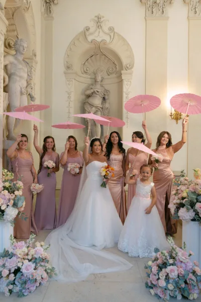 Bridesmaid group photo with bride and bridesmaids holding pink parasols and bouquets in an ornate hall with statues and arched niches