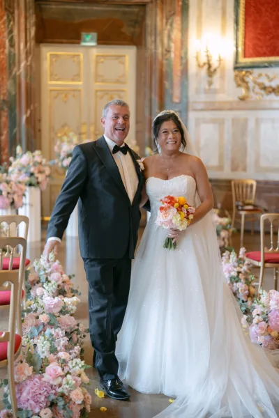 Couple portrait of bride and groom smiling as she holds a bright bouquet, walking an ornate indoor hall aisle with gold chairs