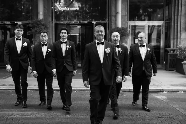 Groomsmen portrait of men in black tuxedos with boutonnieres and bow ties walking together past a glass-door stone entrance on the sidewalk