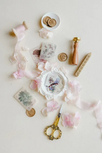 Wax seal stationery and wax seal wedding invitations arranged with pink ribbon, vintage postage stamps, scissors, and petals on a neutral tabletop flat lay