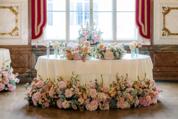 Wedding sweetheart table with sweetheart table flowers, a floral garland of roses, hydrangeas, and eucalyptus beneath tall windows and red drapes