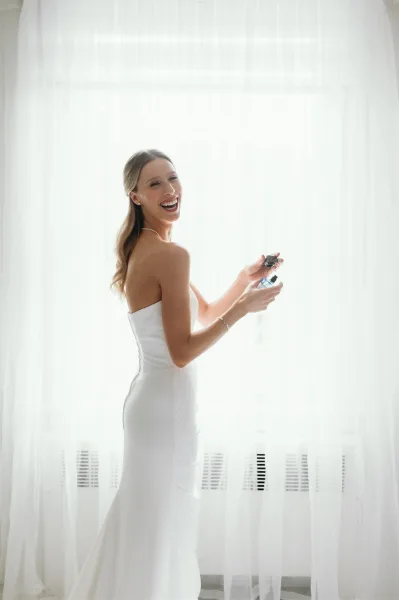 Bridal portrait of a bride with perfume, laughing over her shoulder in a strapless dress and pearl necklace by a window with sheer curtains