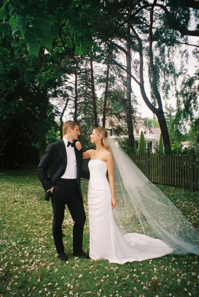 Couple portrait of bride leaning on groom, her cathedral veil train flowing over a garden lawn with tall trees and a wooden fence behind