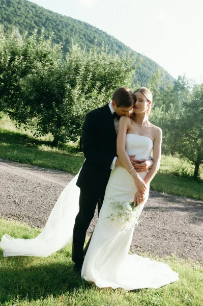 Couple portrait of bride and groom embrace as he kisses her forehead, her strapless gown train and bouquet glowing in sunlit mountains.