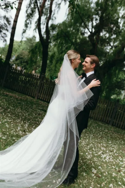 Couple portrait of bride and groom embracing, her long veil trailing over a petal-strewn lawn beside trees and a wooden fence