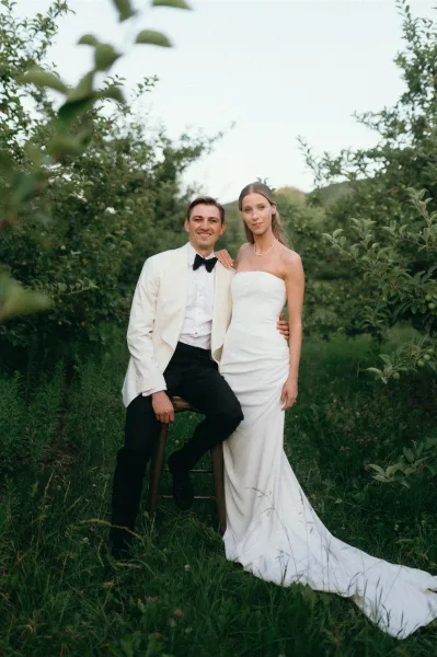 Couple portrait of bride in a strapless gown with long train and groom in white dinner jacket on a stool in an orchard setting