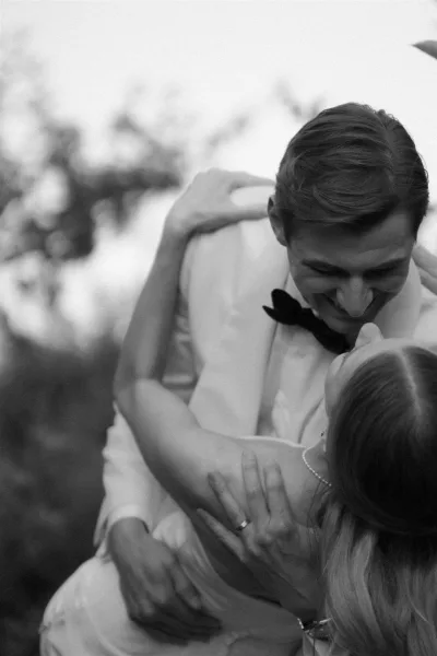 Wedding couple portrait in a black and white wedding photo, groom lifting laughing bride in strapless gown with blurred greenery and sky
