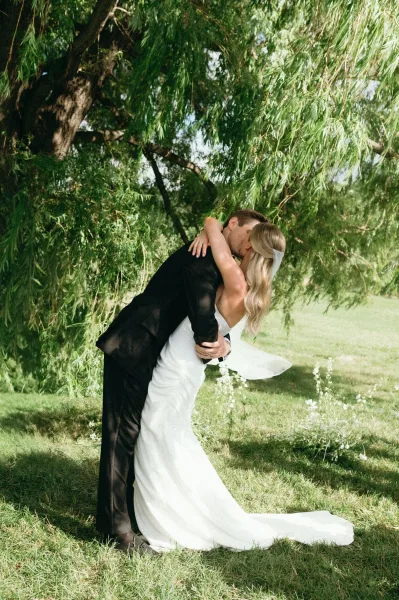 Wedding kiss portrait of the groom dipping the bride as they kiss, her long veil flowing in sunlight beneath a willow tree on a lawn
