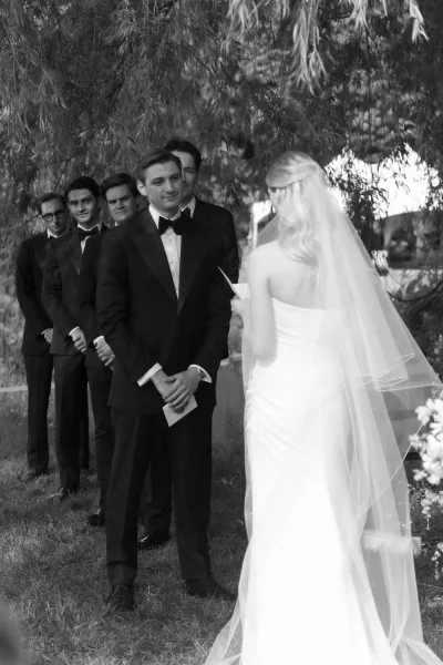 First look moment as bride in long veil approaches groomsmen and groom in black tuxedos, groom reading a note on a tree-lined lawn walkway