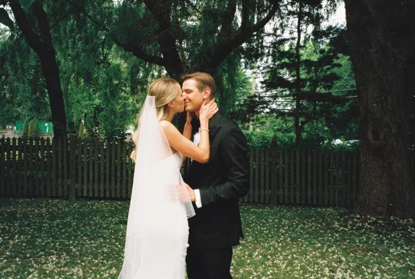 Wedding kiss portrait of bride and groom kissing outdoors, bride holding his face as her veil flows beside trees and a wooden fence