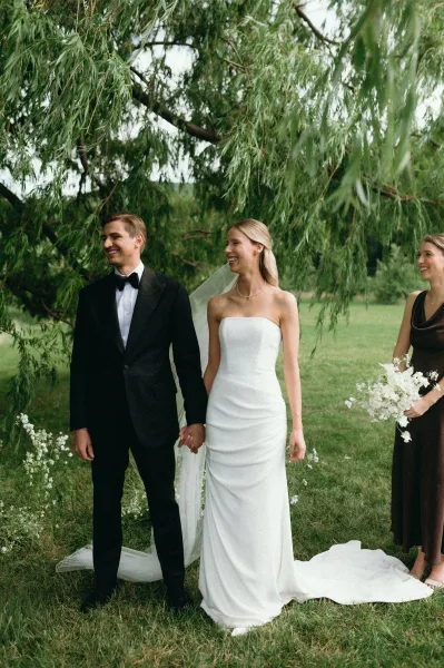Couple portrait of bride and groom holding hands, bride in strapless dress and veil, groom in tuxedo, on a green lawn under trees
