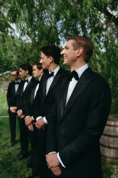 Groomsmen portrait of men lined up in black tuxedos with bow ties and boutonnieres on a green lawn with leafy trees behind