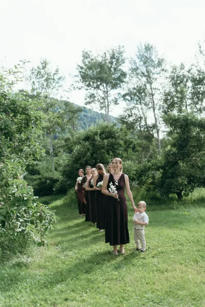Wedding processional with bridesmaids walking down aisle holding white bouquets, ring bearer in suspenders crossing a grassy lawn with mountains beyond
