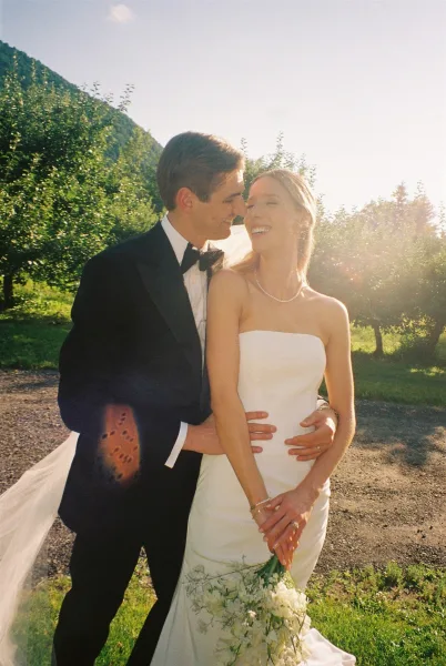 Couple portrait of bride and groom embrace on a mountain hillside, bride in strapless dress and veil with bouquet, sun flare behind trees