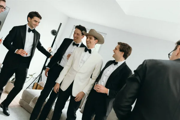 Groomsmen getting ready in tuxedos share a groomsmen toast with whiskey glasses in a living room, one in a white dinner jacket and cowboy hat