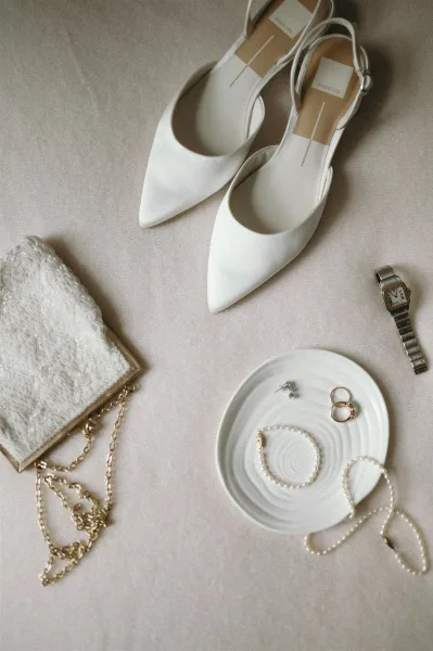 Bridal accessories flat lay with wedding detail flat lay on linen, featuring white pointed toe heels, pearl jewelry, gold rings, and beaded clutch