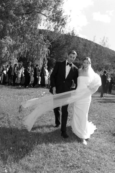 Wedding recessional as bride and groom walking hand in hand, her long veil blowing, guests cheering on a mountain lawn under clouds