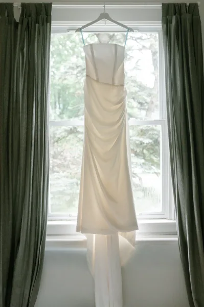 Wedding dress with spaghetti straps hanging on a dress hanger in a bright window, soft curtains framing trees outside in natural light