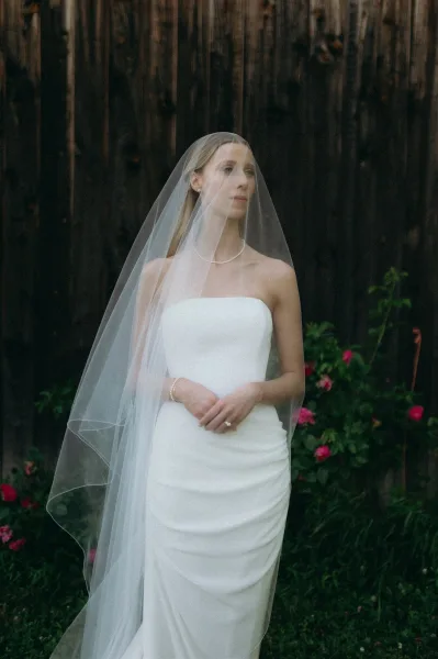 Bridal portrait of a bride in a cathedral veil and strapless wedding dress, posing by a wood fence with garden greenery and pink flowers