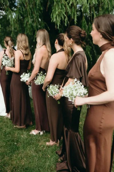 Bridesmaid lineup in brown bridesmaid dresses holding baby’s breath bouquets on a green lawn beneath willow branches outdoors
