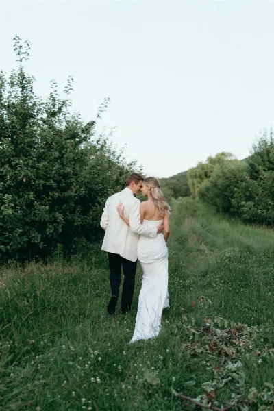 Couple portrait of bride and groom walking away through a wildflower meadow, her strapless dress and his white dinner jacket against rolling hills